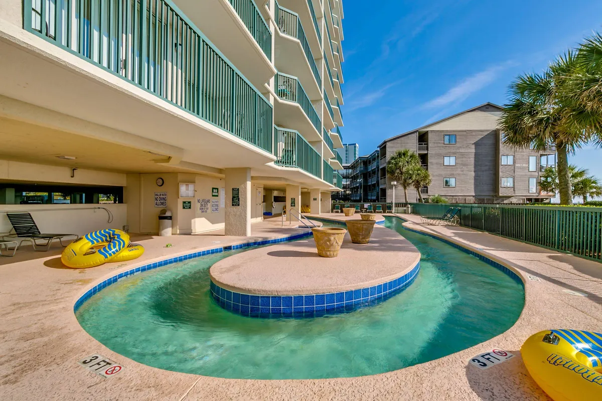 A vibrant pool area featuring a sparkling swimming pool surrounded by poolside lounge chairs, cheerful yellow inner tubes, planters, and balconies overlooking the scene. The atmosphere is lively yet relaxing, suggesting fun for families and groups.