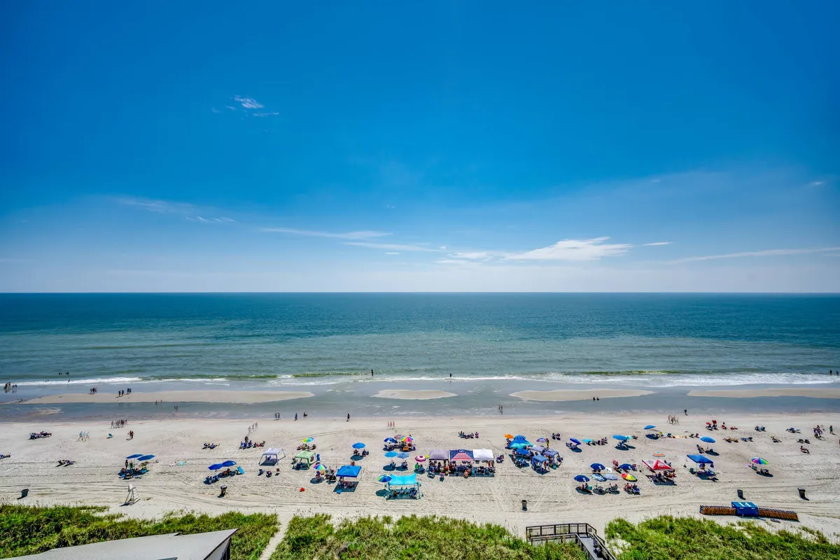 A picturesque beachfront view with colorful umbrellas, gentle waves, sandy shores, and people enjoying a variety of beach activities such as walking, sunbathing, and playing in the sand.