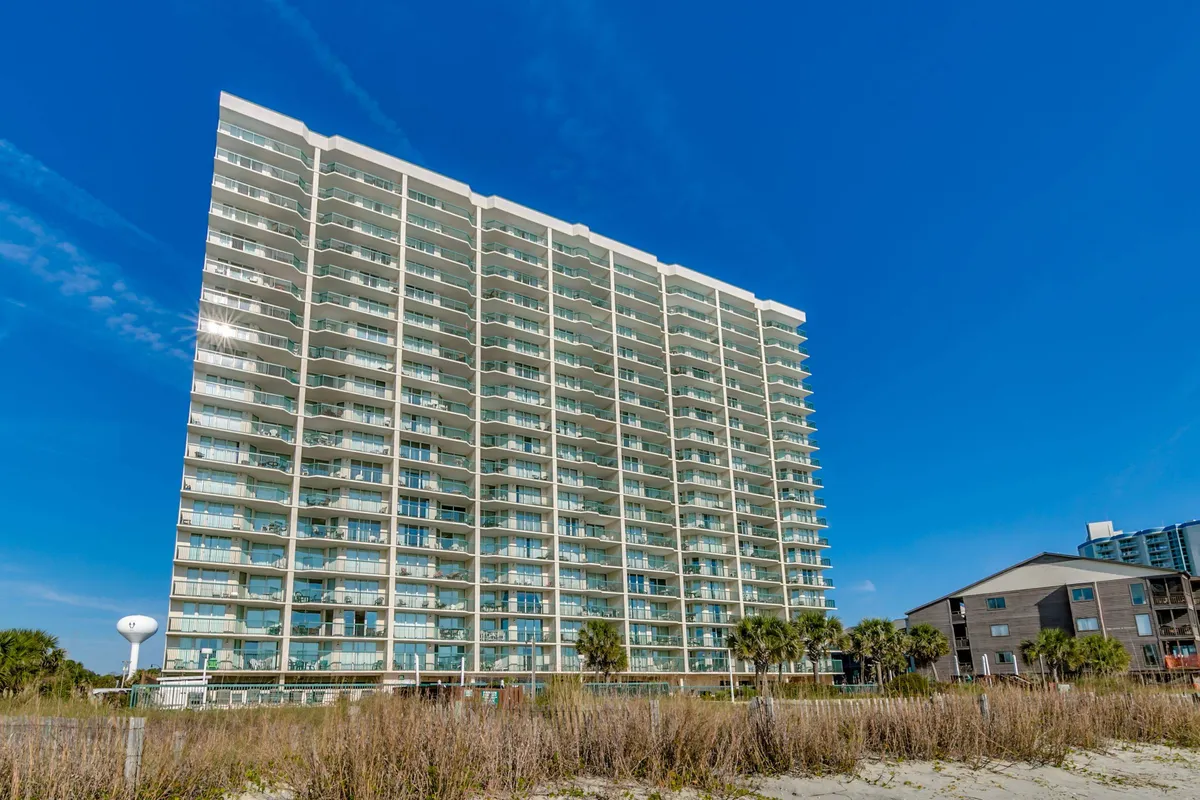 This exterior shot highlights the tall building with multiple balconies, lush greenery, and the surrounding neighborhood. The image conveys a sense of community and provides context for the condo’s setting near golf courses and local attractions.