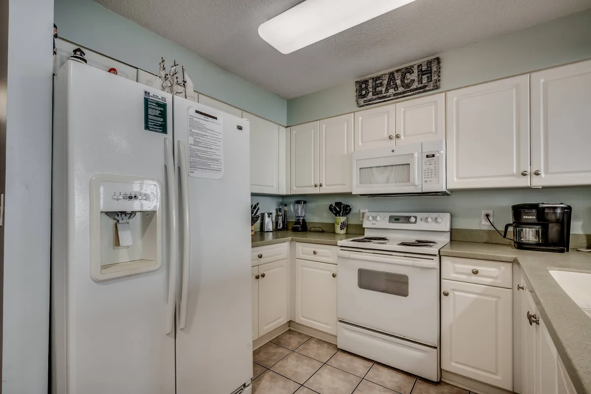 This interior image features a bright, casual kitchen with a white refrigerator, stove and oven, kitchen counter, and a sign that says 'BEACH,' creating a welcoming atmosphere. The space is clearly set up for cooking and gathering, with ample counter space and essential appliances visible.