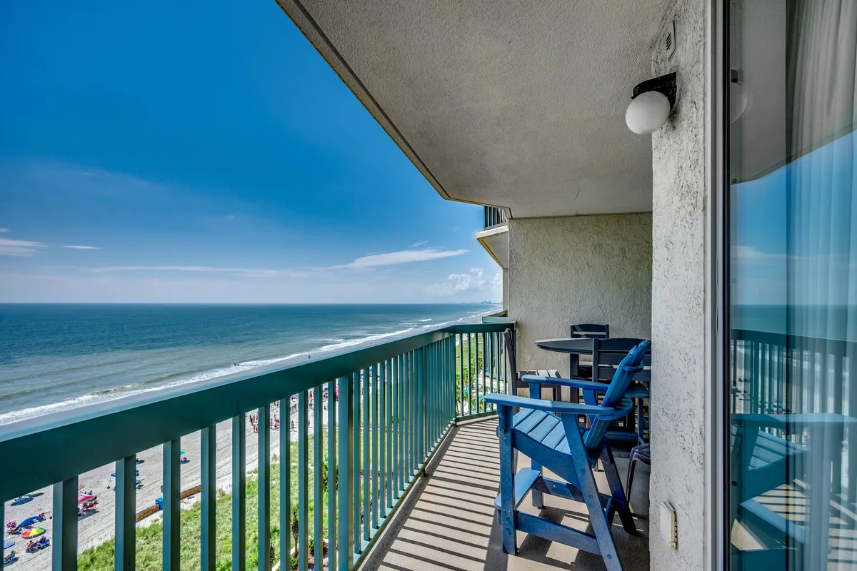 A tranquil balcony scene with comfortable outdoor furniture facing the ocean, under a blue sky. The balcony is the perfect spot for morning coffee or evening conversations, with the sound of waves always present.