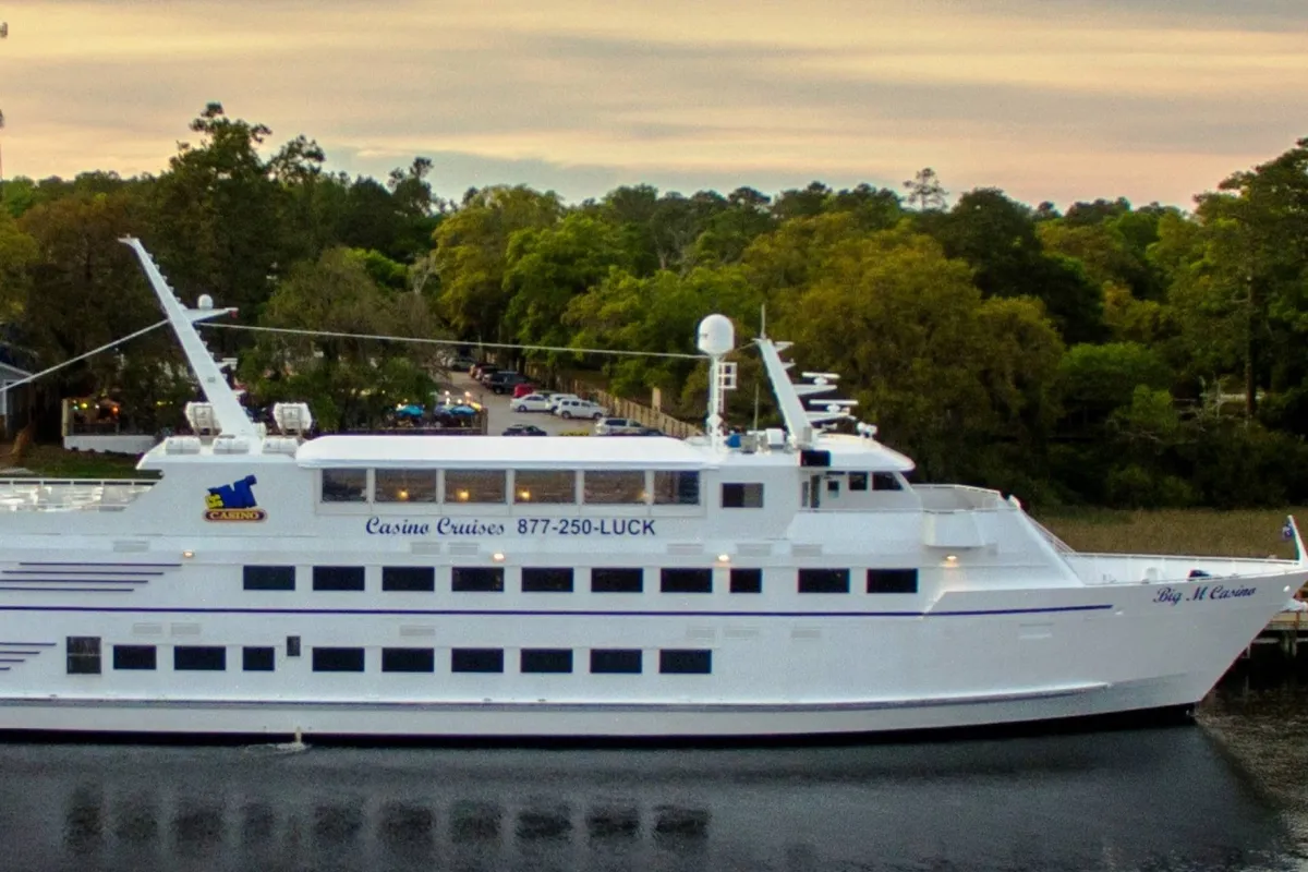 A striking shot of a large white cruise ship branded as a casino, set against natural coastal scenery. The image hints at opportunities for casual dining, socializing, and exciting entertainment aboard the ship, all within the local area.