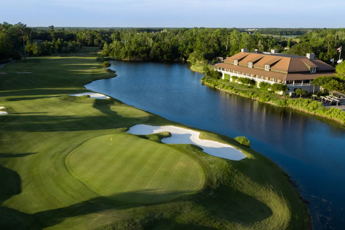 A lush golf course with vibrant green fairways bordering calm blue water, a clubhouse with a brown roof, and sand traps. The peaceful scene highlights guests enjoying golf, picnics, and nature walks.
