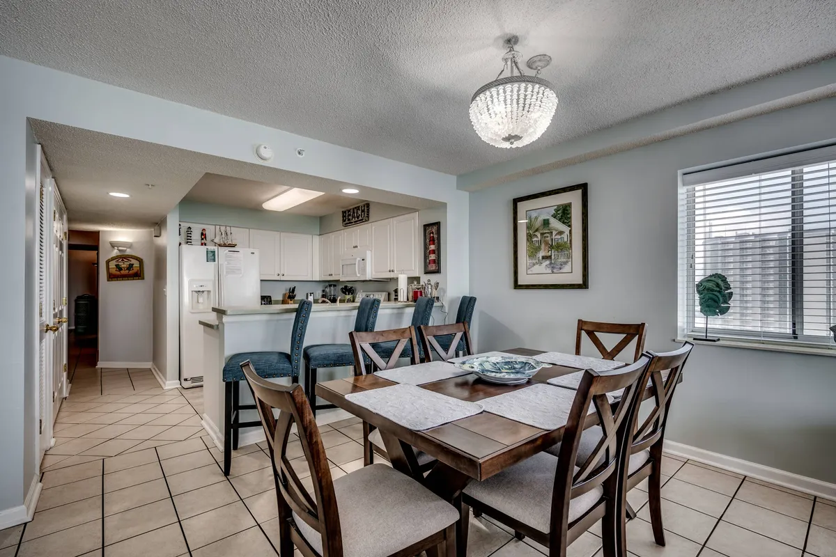 A cheerful dining area with a prominent dining table beneath a chandelier, a nearby kitchen bar area, and a window with blinds that lets in natural light. The setup invites family gatherings and shared meals, perfectly embodying the spirit of communal dining.