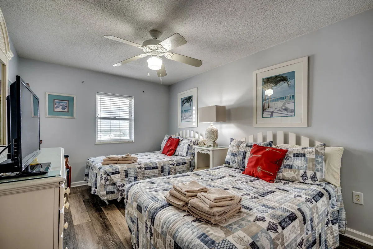 A comfortable bedroom featuring two beds with patterned quilts, colorful decorative pillows, and a television on a dresser. The mood is cozy and welcoming, ideal for children or friends sharing a room.