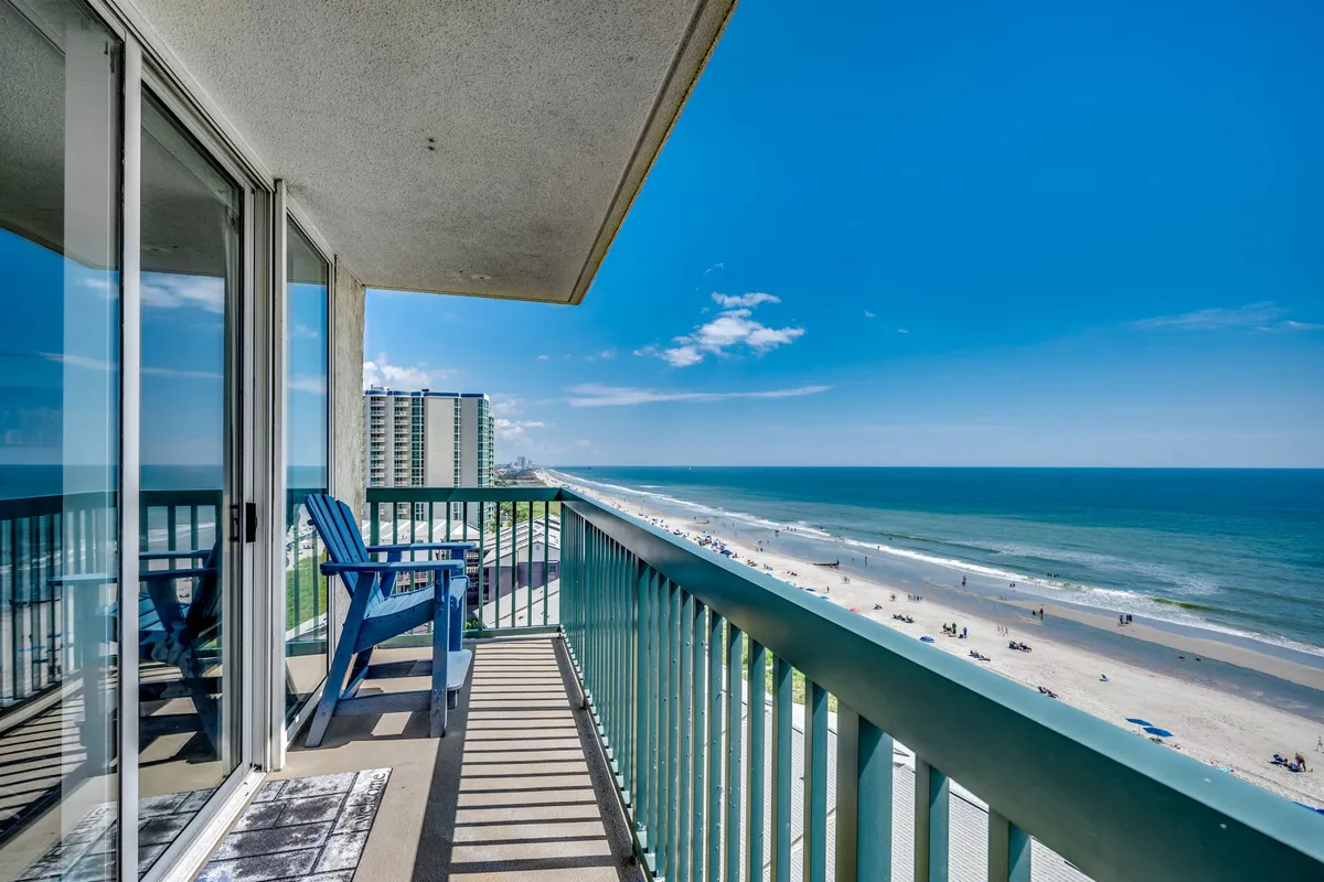 A tranquil scene from a balcony overlooking the ocean and sandy beach, with comfortable blue chairs perfect for relaxing. The wide-open view highlights the proximity to the water and the prime oceanfront location.