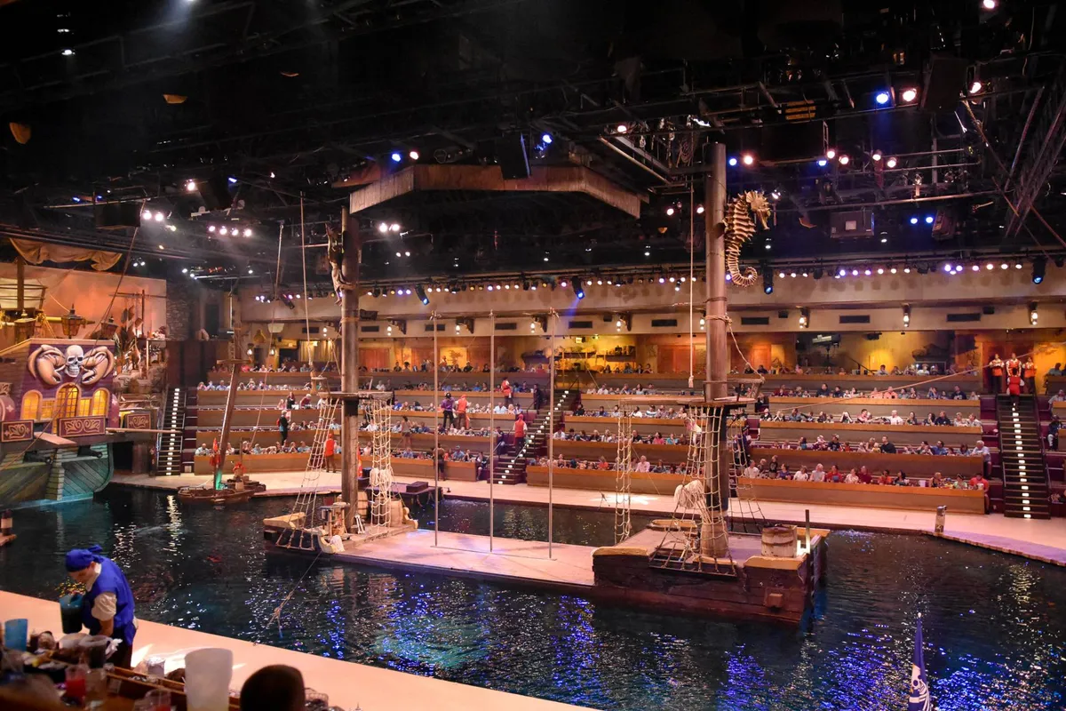 A dynamic interior scene featuring a central stage with a water feature, elaborately decorated pirate ship set design, and spacious audience seating. Stage lighting and aquatic elements hint at high-energy performances and immersive experiences.