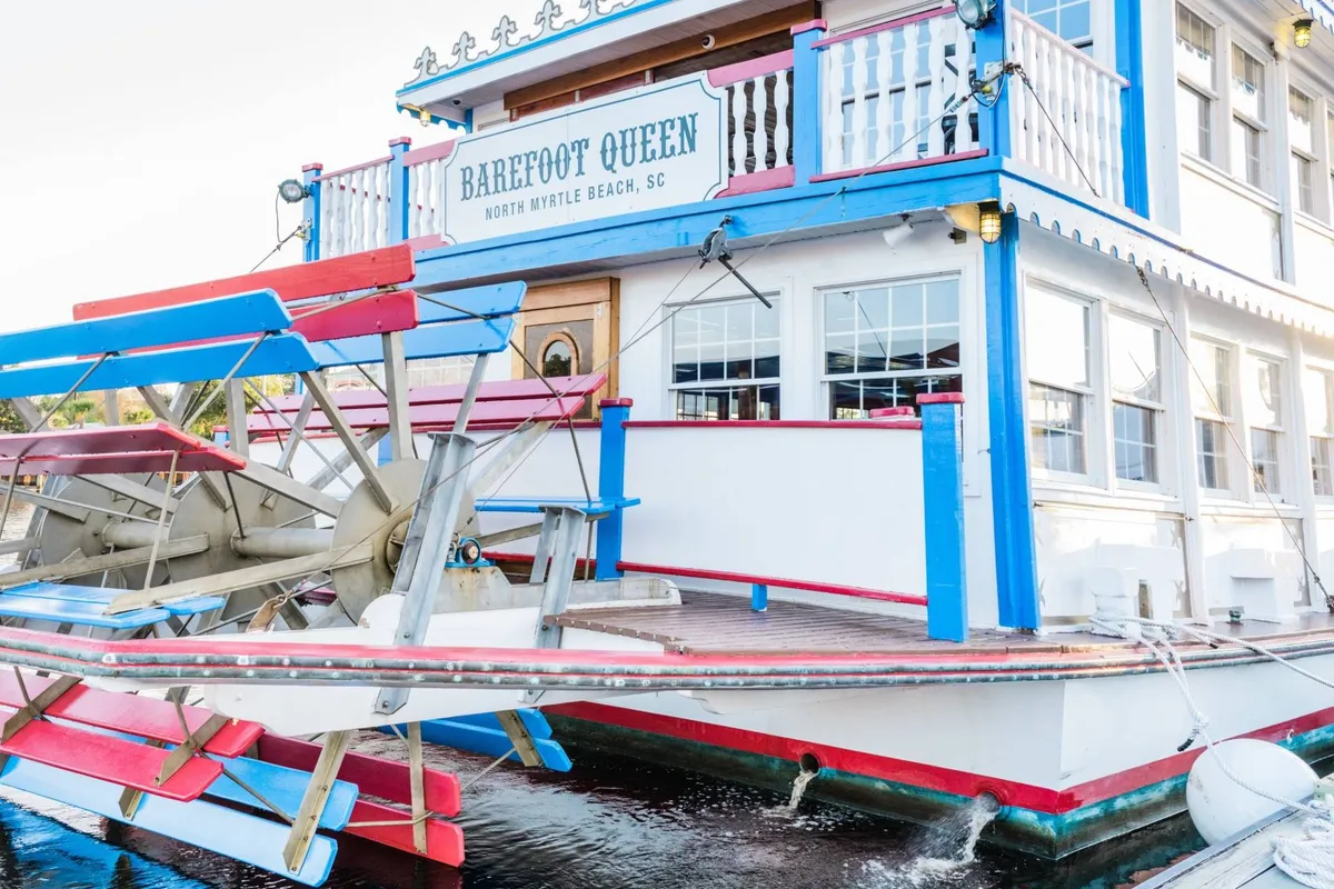 A lively exterior shot of the Barefoot Queen riverboat, showcasing the paddle wheel, ornate signage, and colorful trim. The festive atmosphere suggests social gatherings, themed dining, and river cruising adventures.